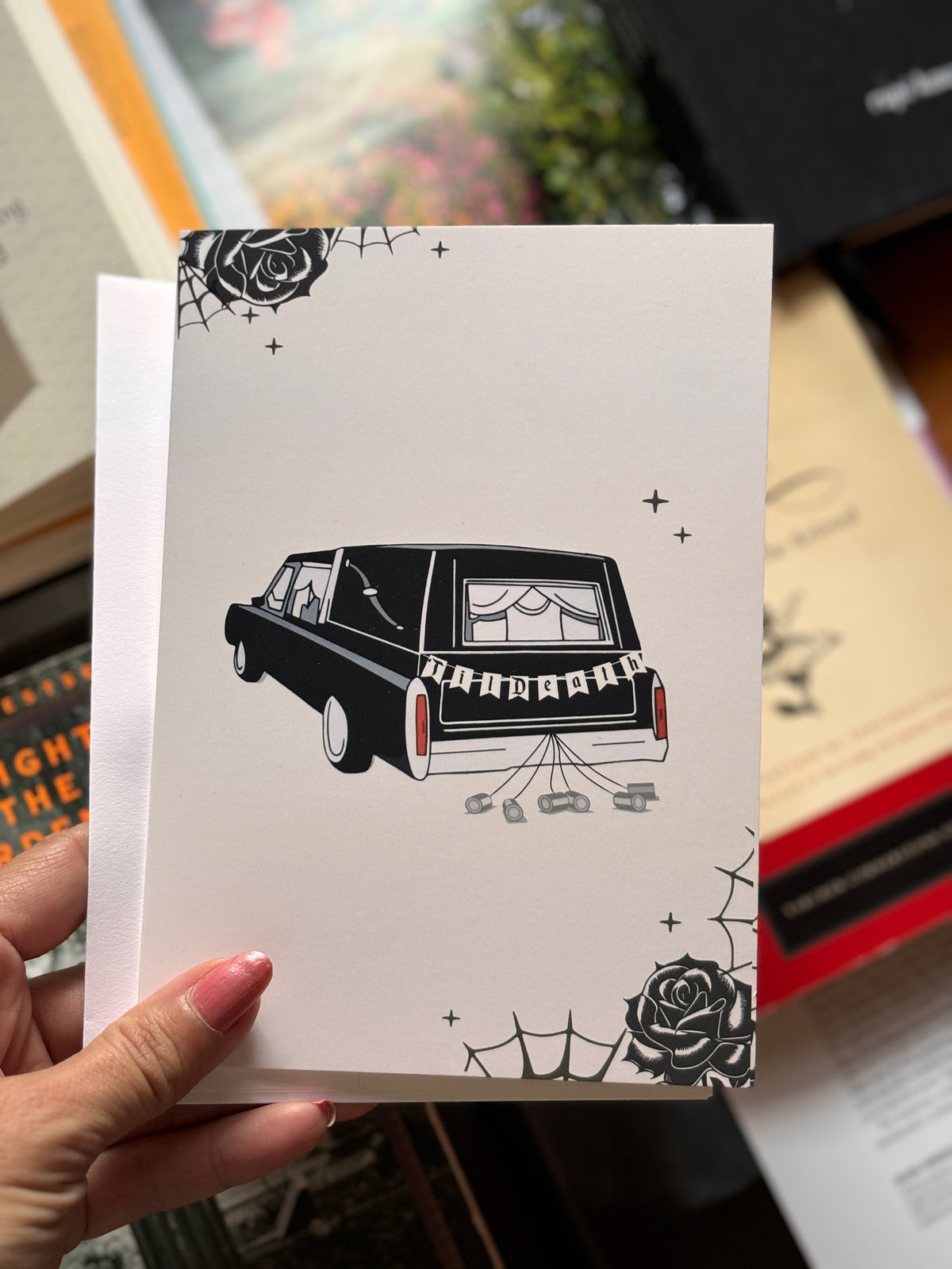 A cream-colored greeting card featuring a black-and-white illustration of a hearse with 'TIl Death' on the back, decorated with spiderwebs and roses.