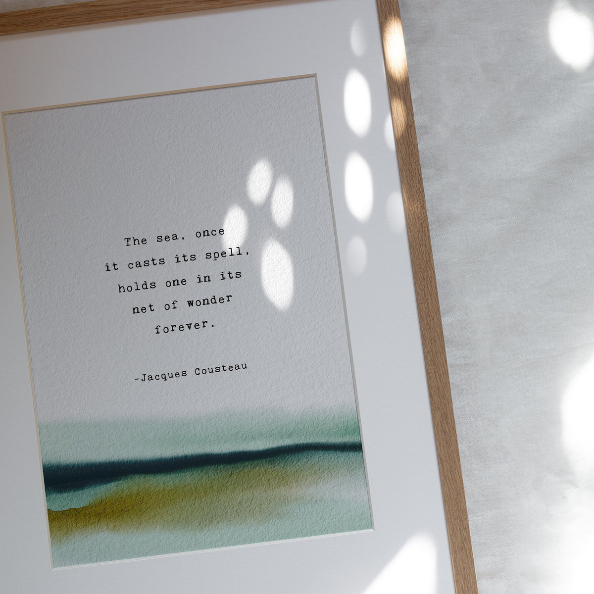 An art print featuring a quote by Jacques Cousteau on a soft watercolor background which says "the sea, once it casts its spell, holds one in its net of wonder forever"