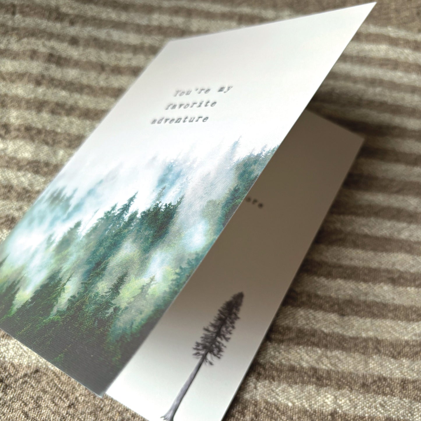 A folded greeting card with a watercolor forest design and the printed phrase 'You're my favorite adventure' lies on a textured brown and beige striped fabric surface.