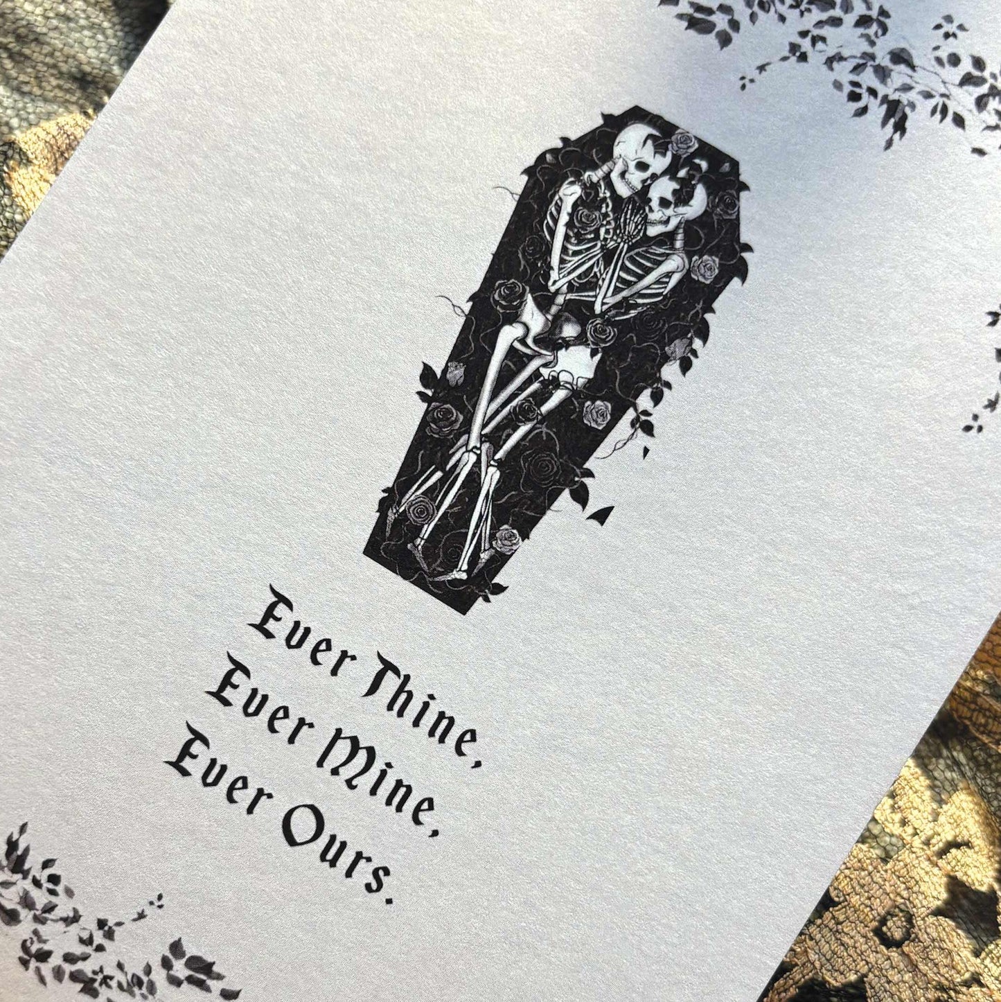 A close-up of a white printed card or paper featuring a black coffin-shaped illustration with two skeletons and the gothic text 'Ever Thine, Ever Mine, Ever Ours.' 