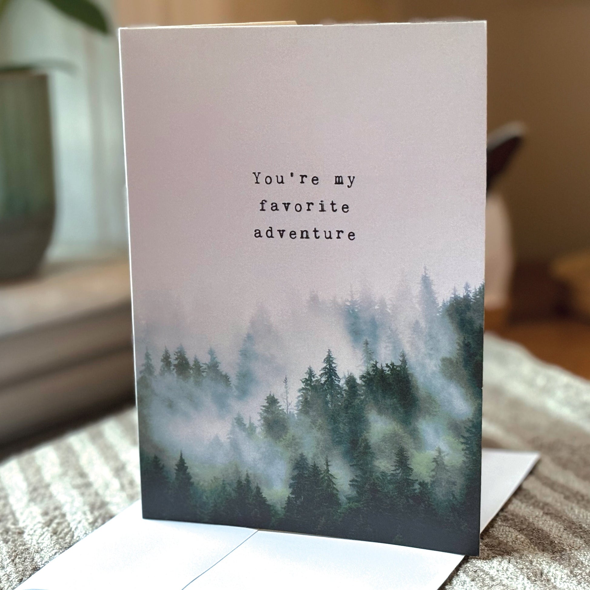 A vertical greeting card with a misty pine forest illustration and the printed phrase 'You're my favorite adventure' in black typewriter-style text. 