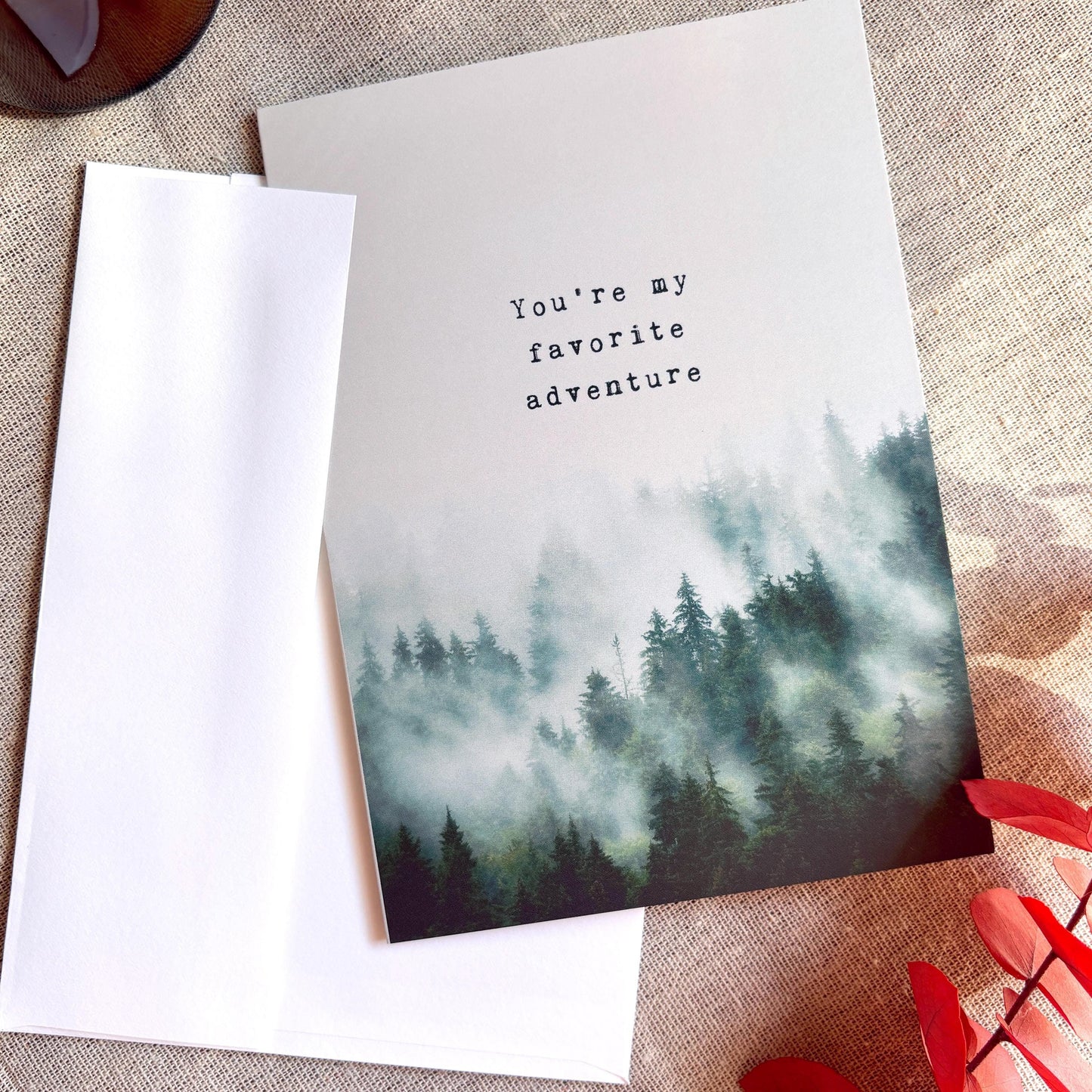 A  greeting card with a misty pine forest illustration and the text 'You're my favorite adventure,' shown with a white envelope on a textured beige fabric background.