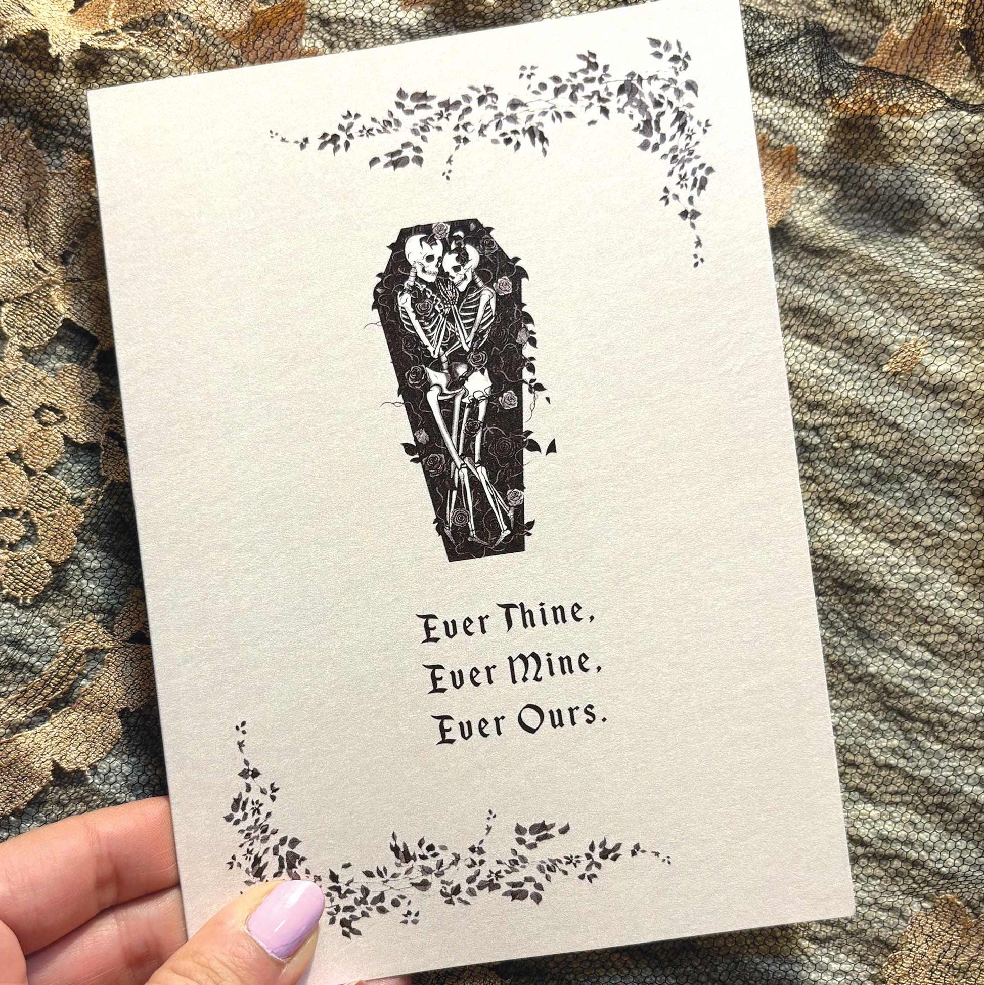 A greeting card featuring a black ink illustration of a skeleton in a coffin, surrounded by floral vines and the text 'Ever Thine. Ever Mine. Ever Ours.