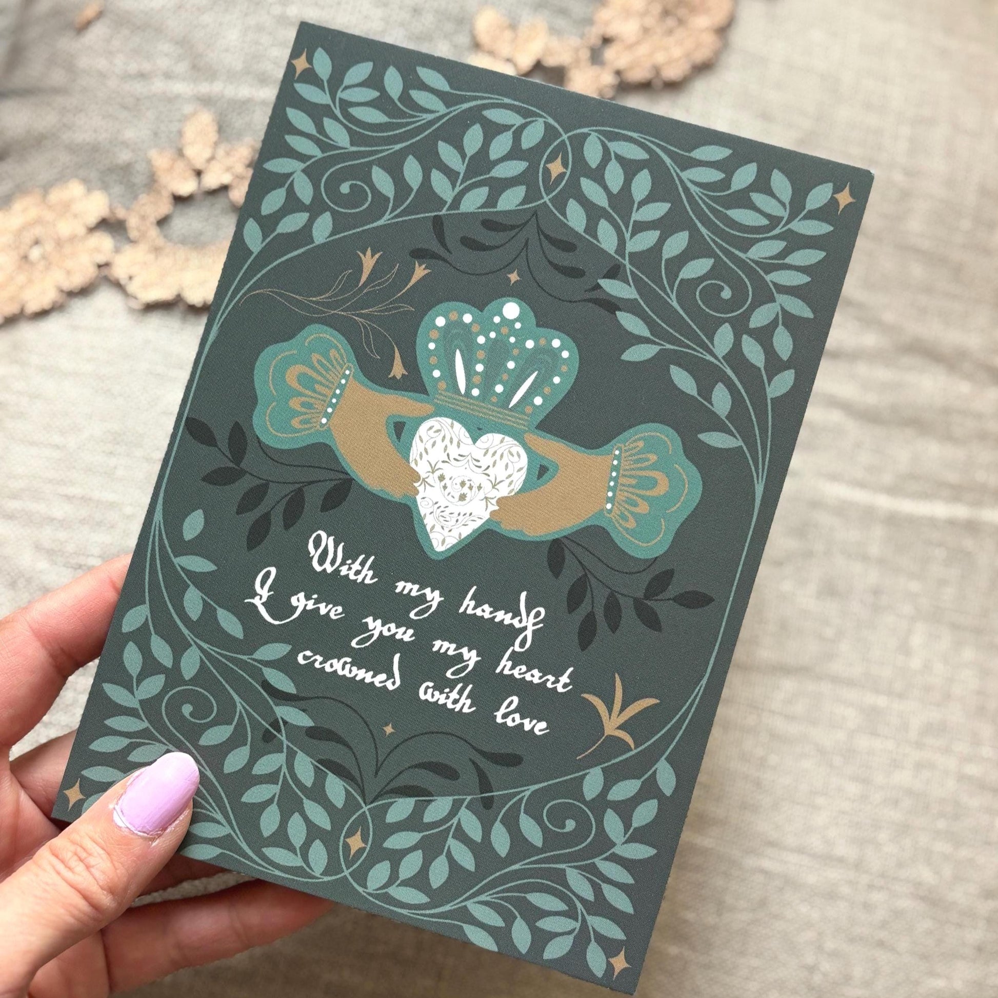 a dark teal greeting card featuring an Irish Claddagh-style heart design with a white heart, gold and teal hands, and white cursive text reading, 'With my hands I give you my heart, crowned with love.' The background is a neutral textured fabric with dried flowers.