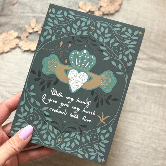 a dark teal greeting card featuring an Irish Claddagh-style heart design with a white heart, gold and teal hands, and white cursive text reading, 'With my hands I give you my heart, crowned with love.' The background is a neutral textured fabric with dried flowers.