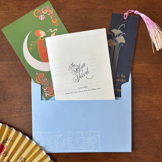 A flat-lay of a stationery suite featuring a light blue envelope with a white 'The Blue Velvet' , a green card with an orange crescent moon illustration, a dark navy bookmark with gold floral line art and a pink tassel