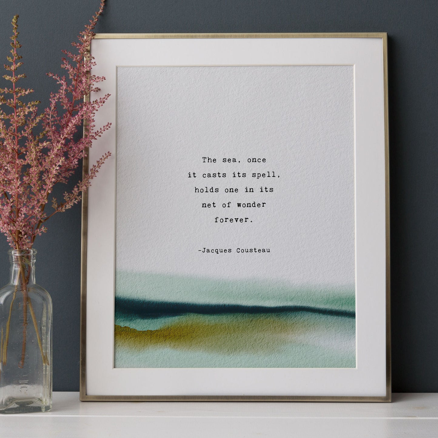 A vertical art print with a watercolor landscape at the bottom and a printed quote by Jacques Cousteau in the center, which says "the sea, once it casts it spell, holds one in its net of wonder forever"