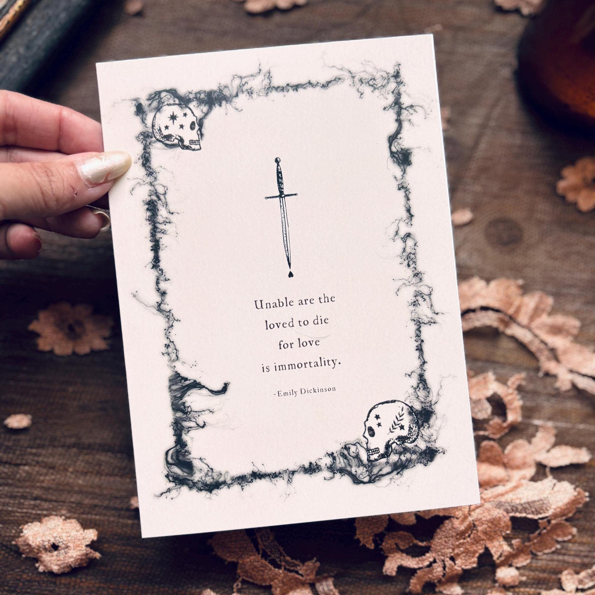 A white greeting card with a black gothic illustration of a skull and dagger, featuring a quote by Emily Dickinson which says "Unable are the loved to die for love is immortality"