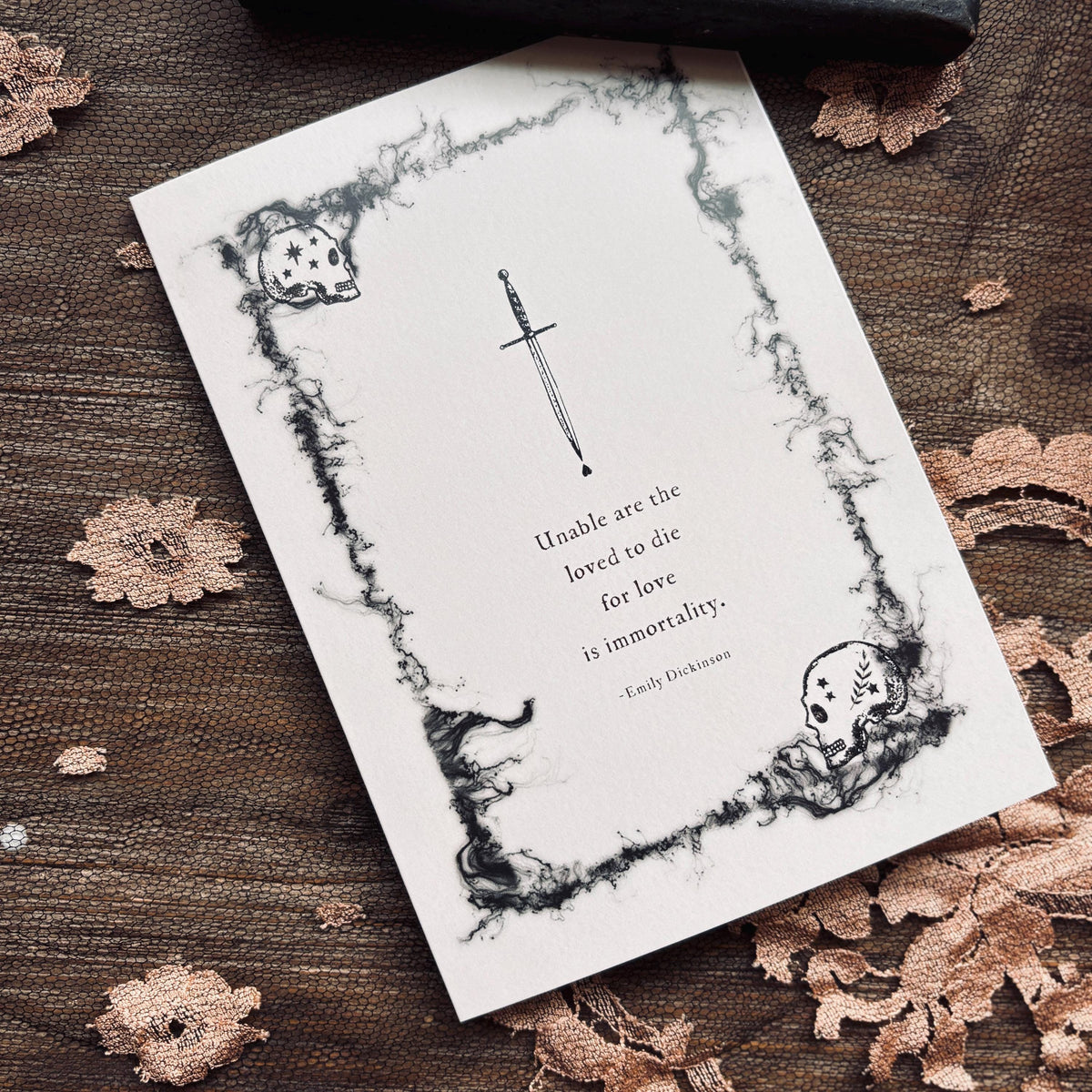 A white greeting card with a black gothic illustration of a skull and dagger, featuring a quote by Emily Dickinson which says "Unable are the loved to die for love is immortality"