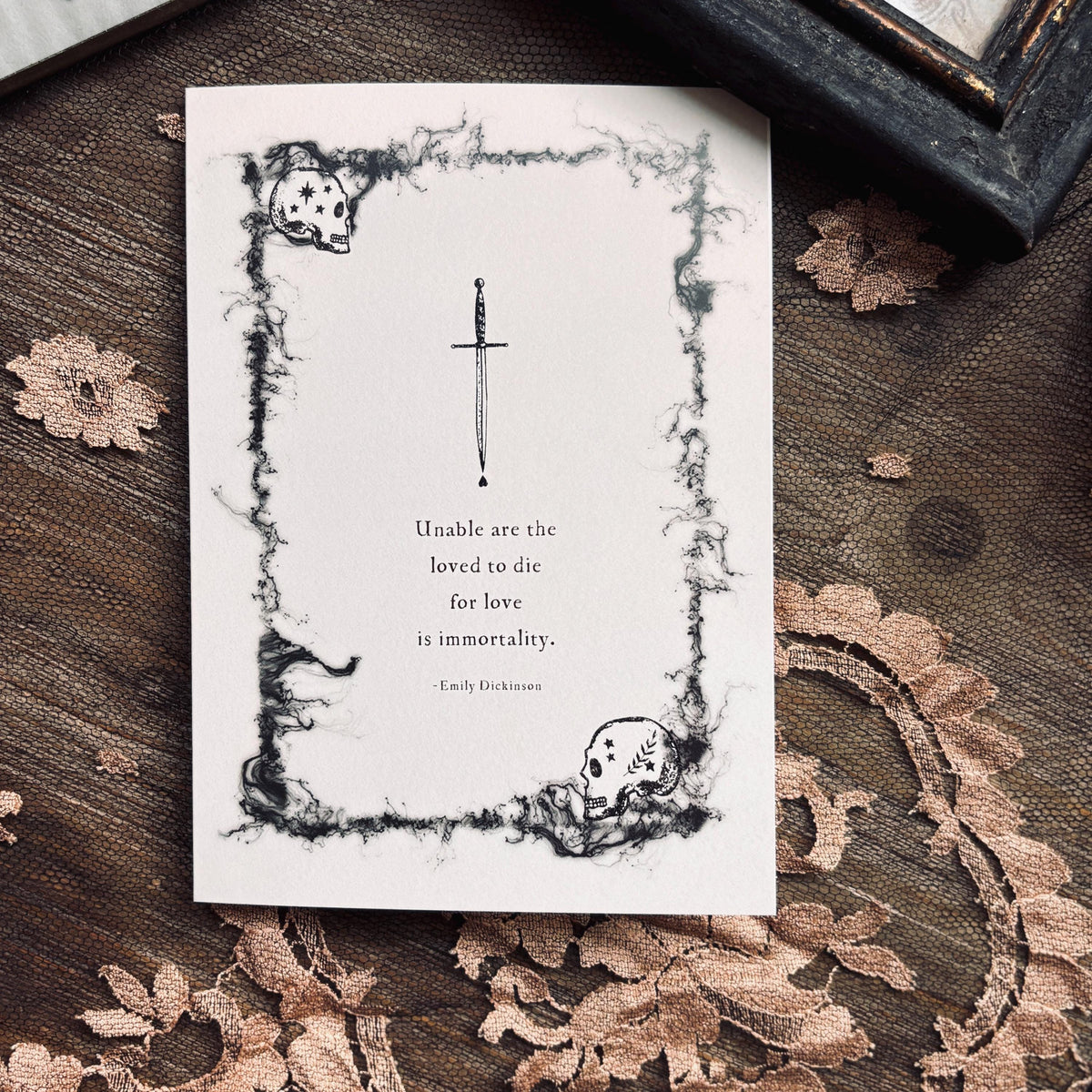 A white greeting card with a black gothic illustration of a skull and dagger, featuring a quote by Emily Dickinson which says "Unable are the loved to die for love is immortality"