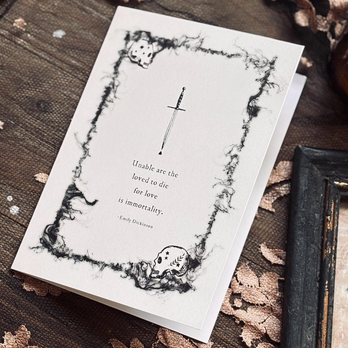 A white greeting card with a black gothic illustration of a skull and dagger, featuring a quote by Emily Dickinson which says "Unable are the loved to die for love is immortality"