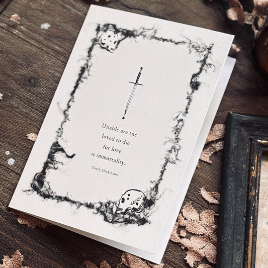 A white greeting card with a black gothic illustration of a skull and dagger, featuring a quote by Emily Dickinson which says "Unable are the loved to die for love is immortality"