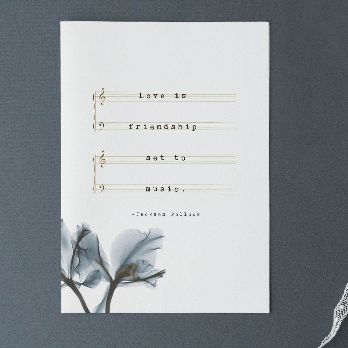 A typographic art print featuring the quote 'Love is friendship set to music.' on musical staff lines, with a small blue-gray floral illustration in the lower left and the text '-Jackson Pollock' near the bottom.