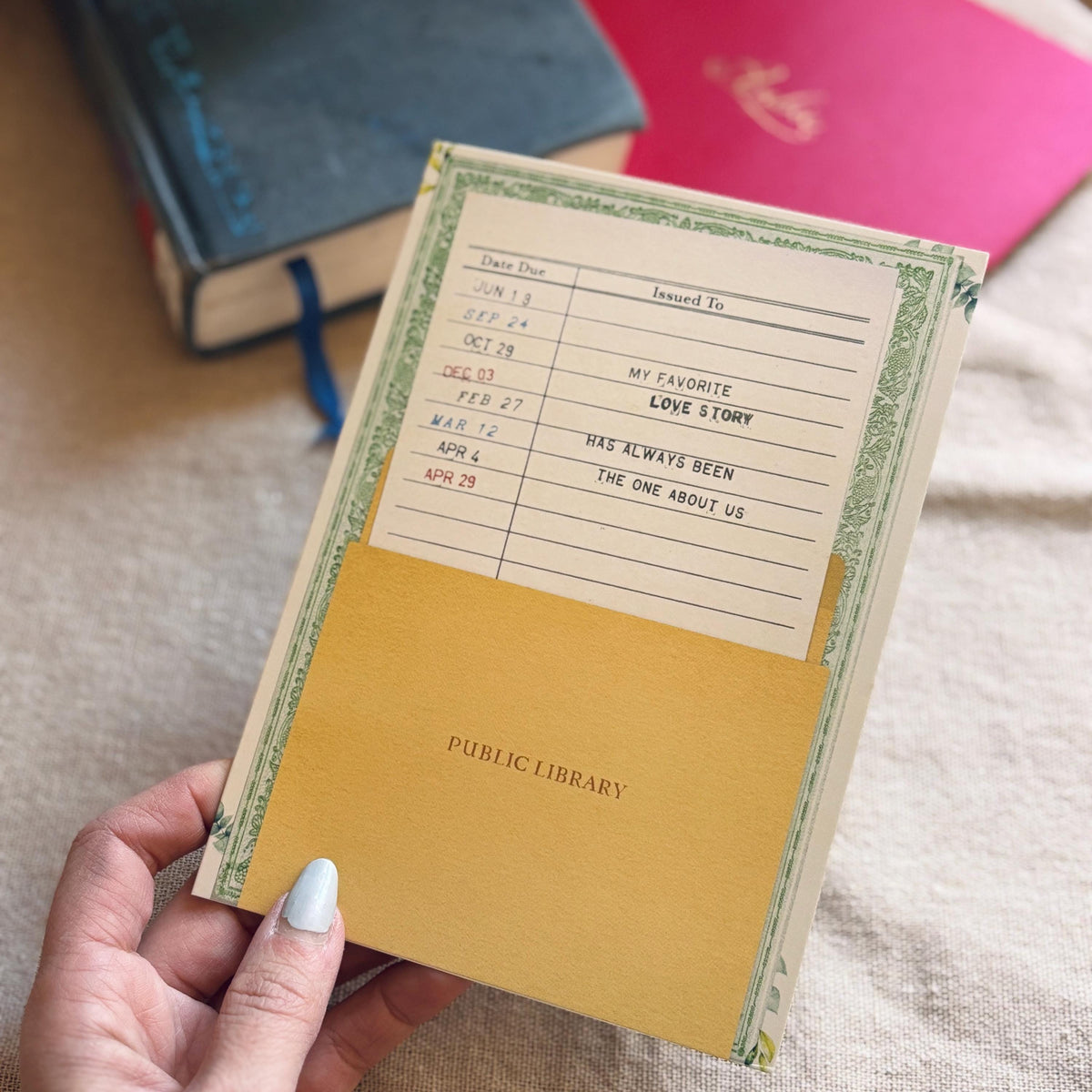 A vintage-style public library greeting card The cream-colored card features a green floral border, a date due section, and the message "my favorite love story has always been the one about us"