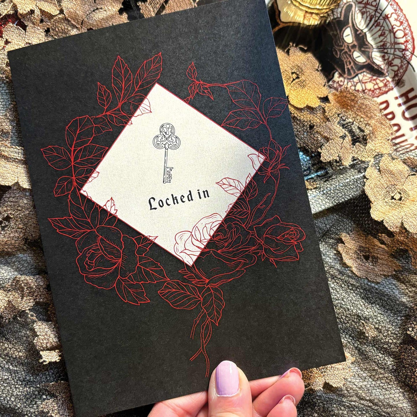 A black greeting card featuring  a key illustration and the words 'Locked in,' surrounded by red rose line drawings.