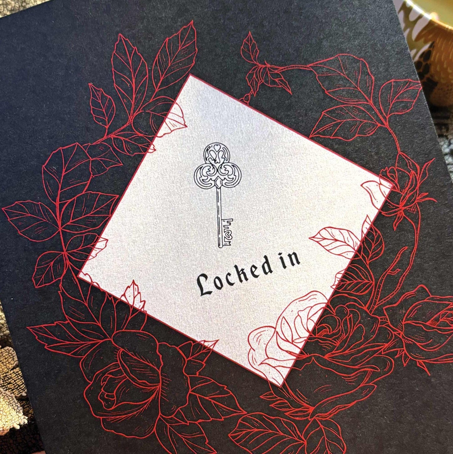 A black greeting card featuring  a key illustration and the words 'Locked in,' surrounded by red rose line drawings.