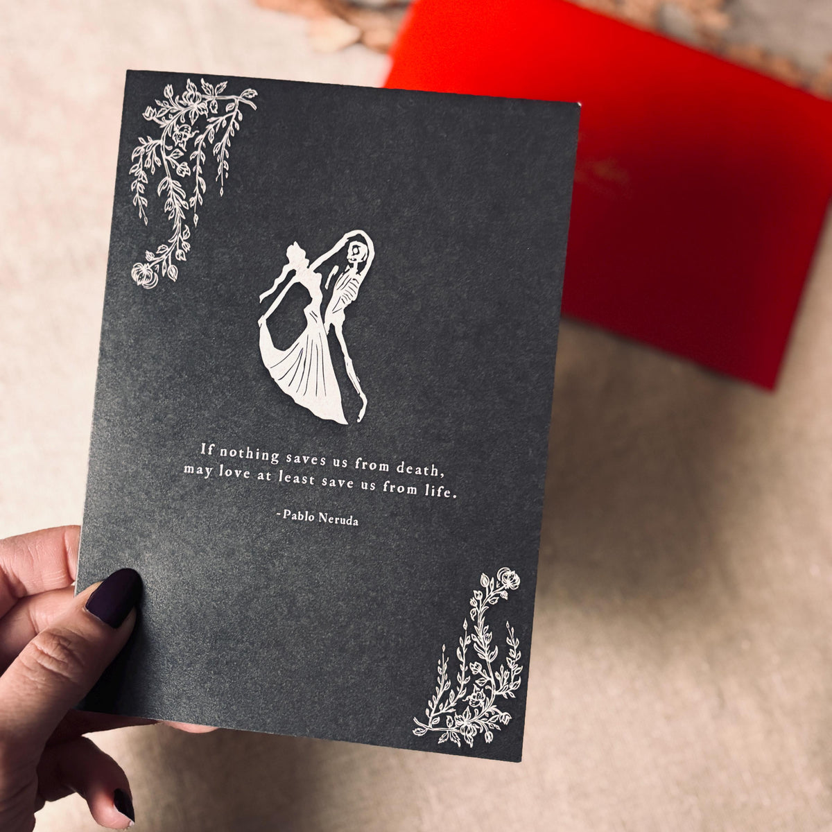 A black greeting card featuring a white illustration of a dancing skeleton couple, a quote by Pablo Neruda, and decorative corner floral motifs. it says "If nothing saves us from death, may love at least save us from life."