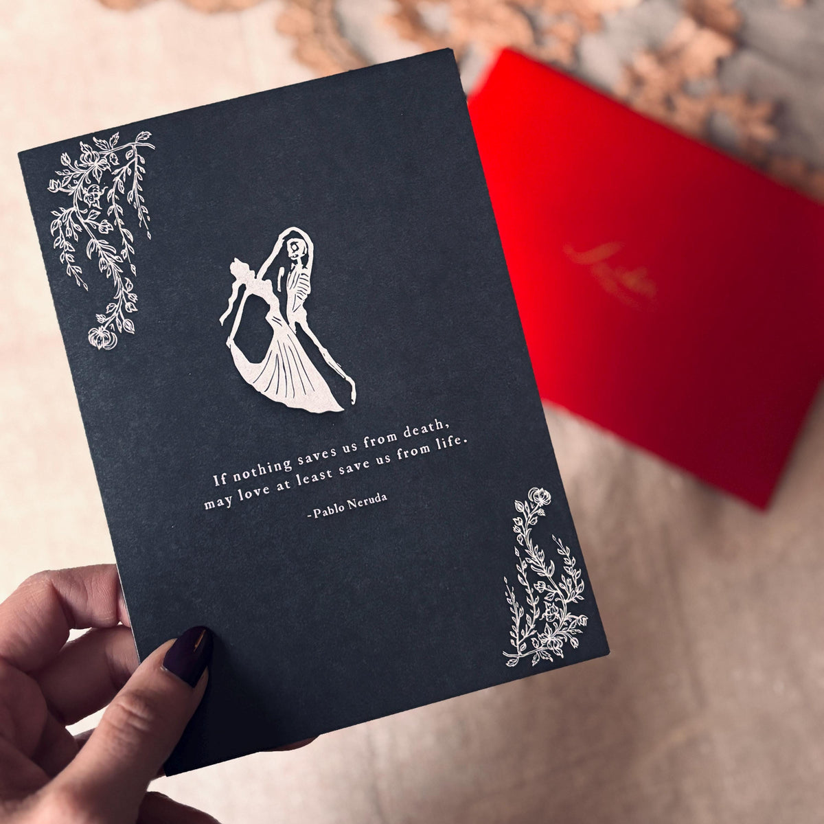 A black greeting card featuring a white illustration of a dancing skeleton couple, a quote by Pablo Neruda, and decorative corner floral motifs. it says "If nothing saves us from death, may love at least save us from life."