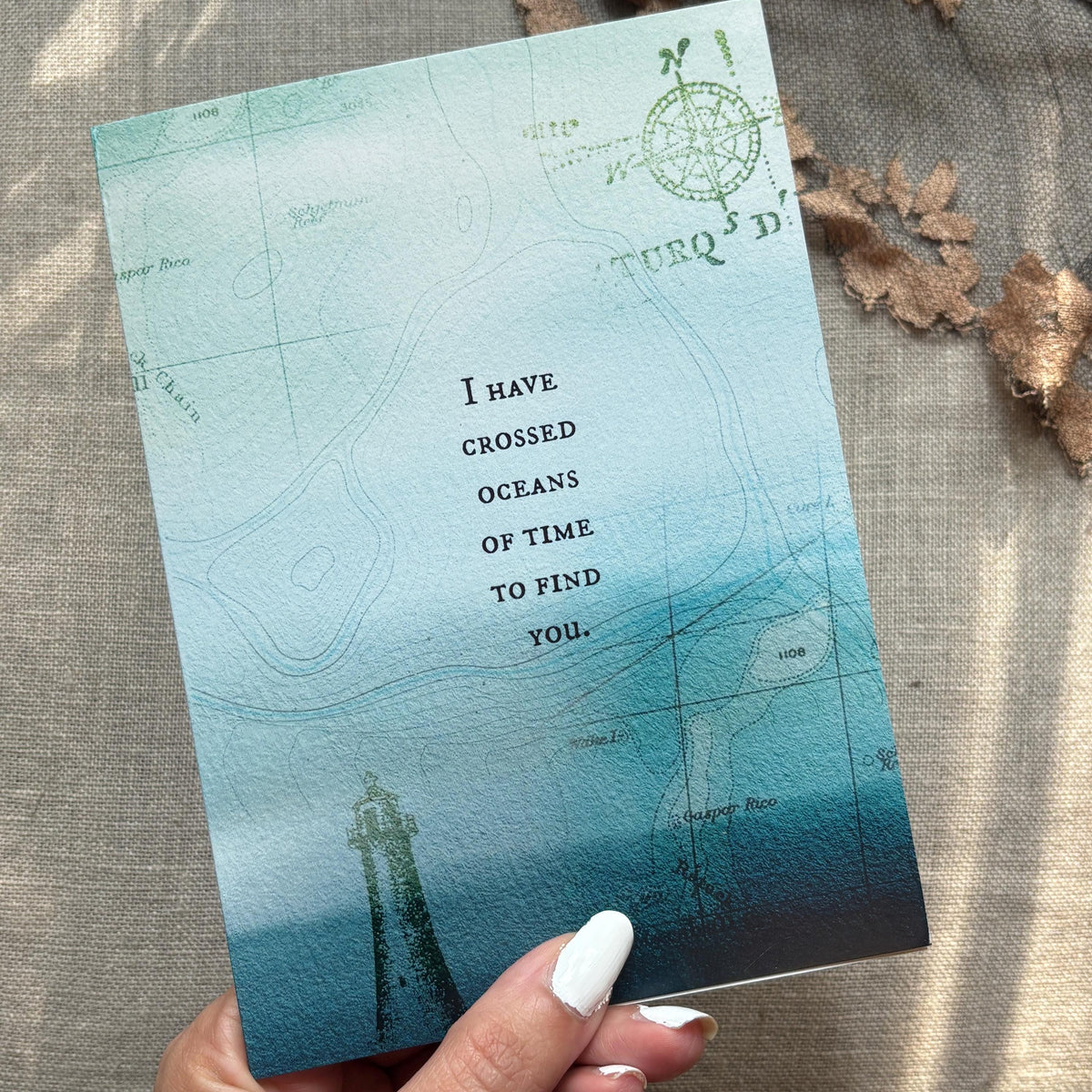 A greeting card featuring a lighthouse, a compass rose, and a blue-to-teal gradient with the text 'I have crossed oceans of time to find you' 