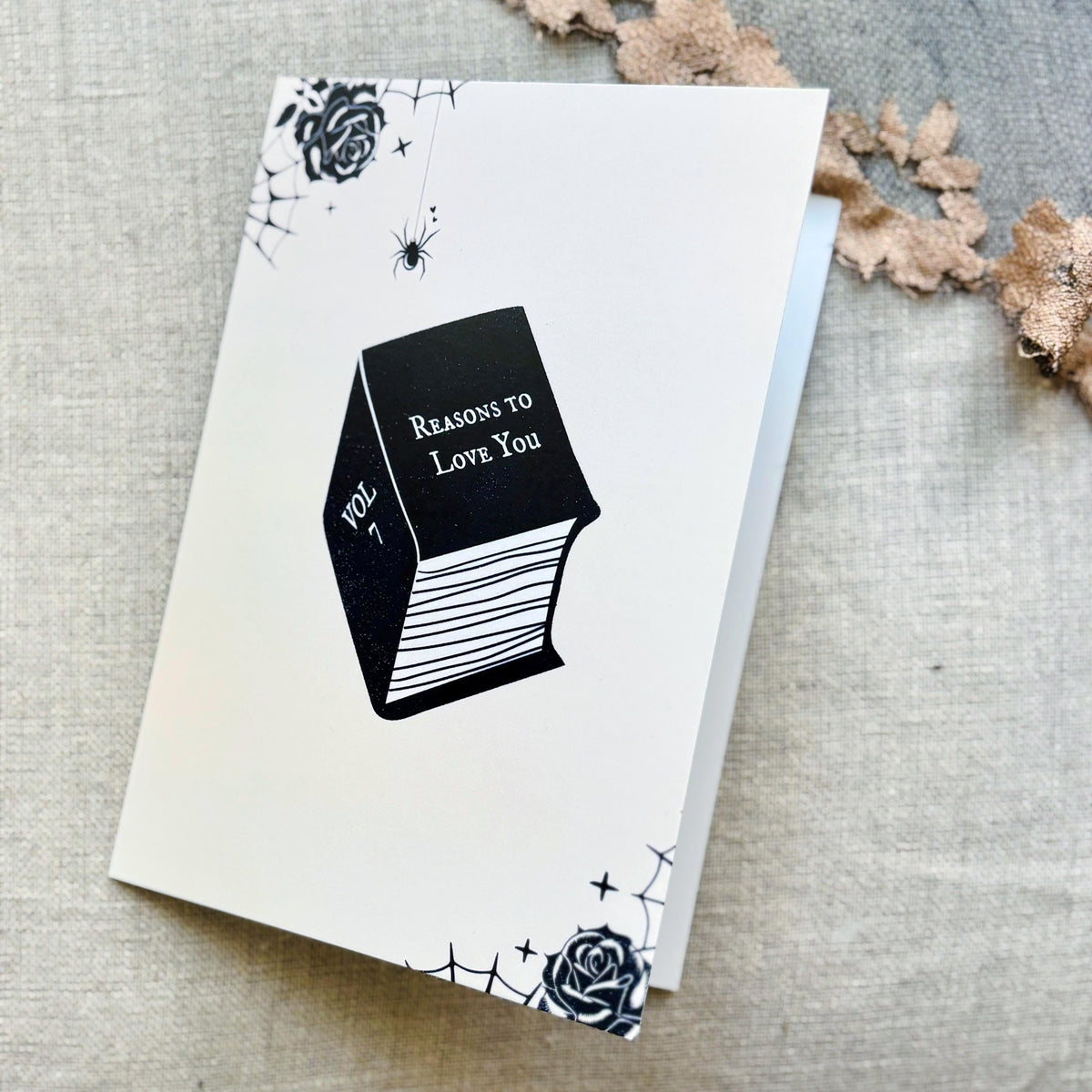 A white greeting card featuring a black illustration of a book titled 'Reasons to Love You' with spiderwebs, roses, and stars