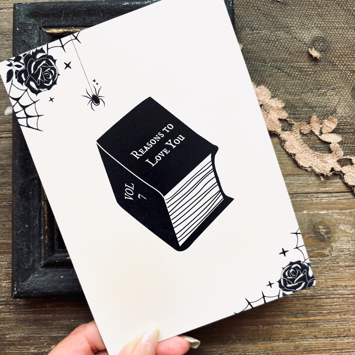 A white greeting card featuring a black illustration of a book titled 'Reasons to Love You' with spiderwebs, roses, and stars