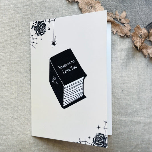 A white greeting card featuring a black illustration of a book titled 'Reasons to Love You' with spiderwebs, roses, and stars