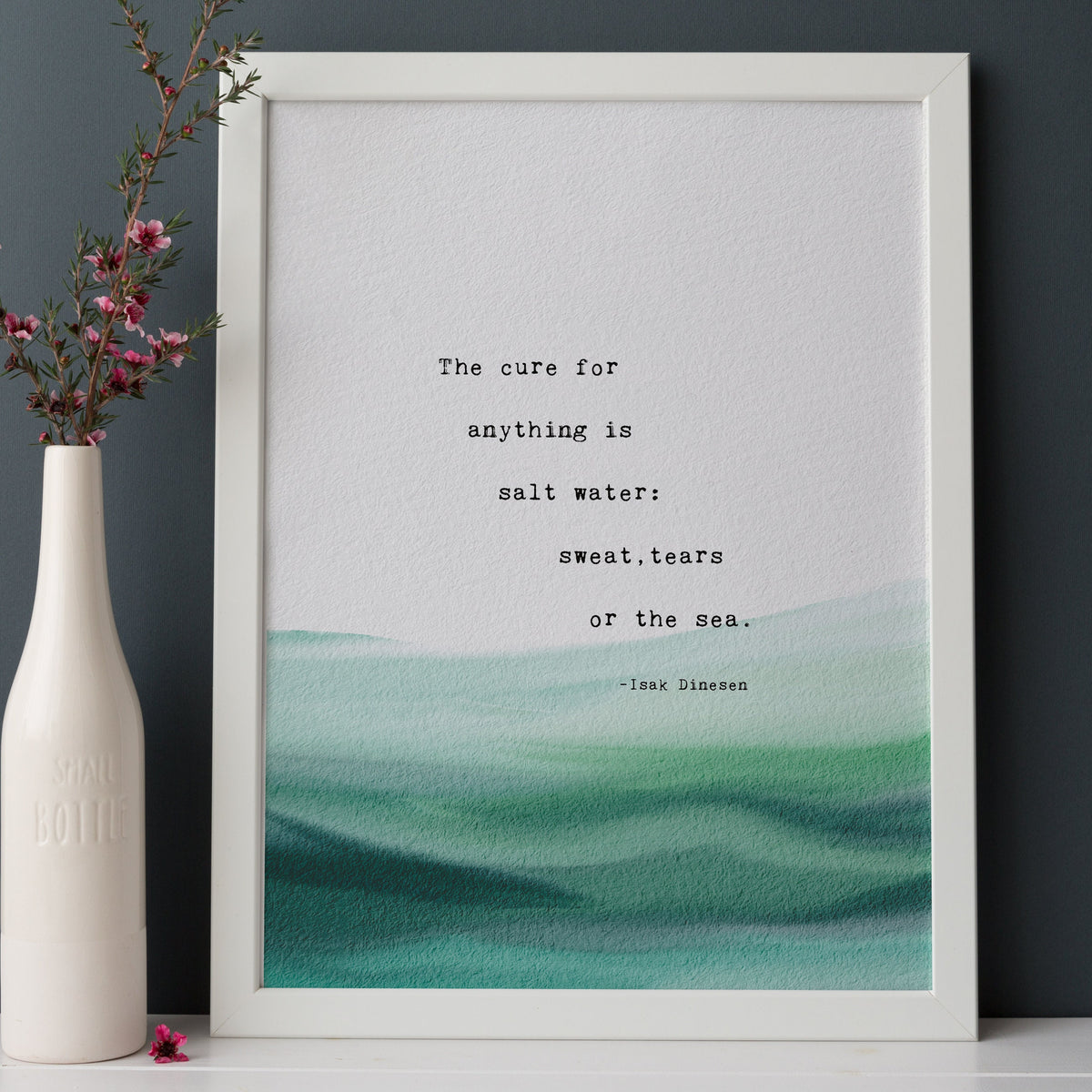 A typographic art print featuring a quote by Isaac Dinesen over a watercolor ocean wave design in shades of teal and green on a white background. "The cure for anything is salt water: sweat, tears or the sea"