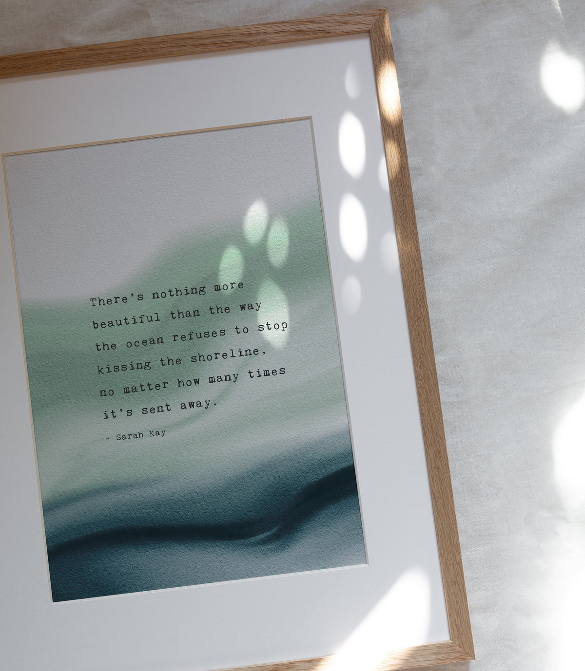 A vertical art print featuring a quote by Sarah Ray over an abstract watercolor background of soft green and deep teal. The quote says "there's nothing more beautiful than the way the ocean refuses to stop kissing the shoreline, no matter how many times it's sent away"