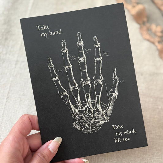A black greeting card featuring a white anatomical illustration of a hand skeleton with the text 'Take my hand' and 'Take my whole life too'.