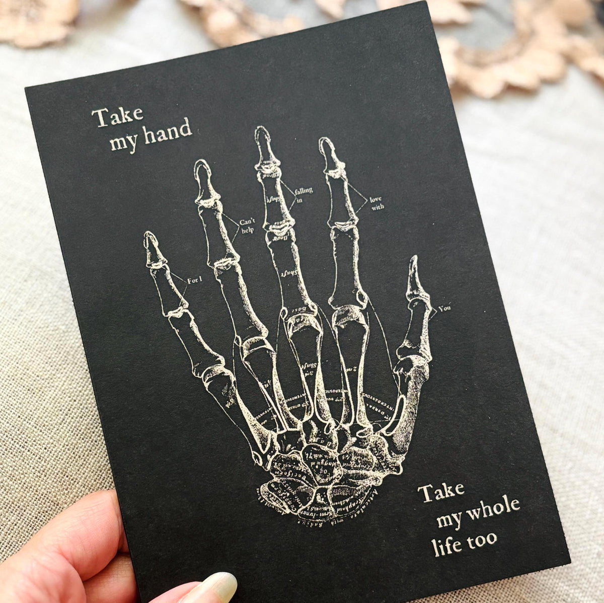 A black greeting card featuring a white anatomical illustration of a hand skeleton with the text 'Take my hand' and 'Take my whole life too'.