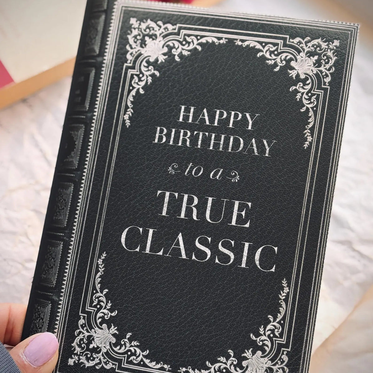 A birthday card with a black faux-leather texture with ornate silver decorative corners and the text 'HAPPY BIRTHDAY to a TRUE CLASSIC' on the front.