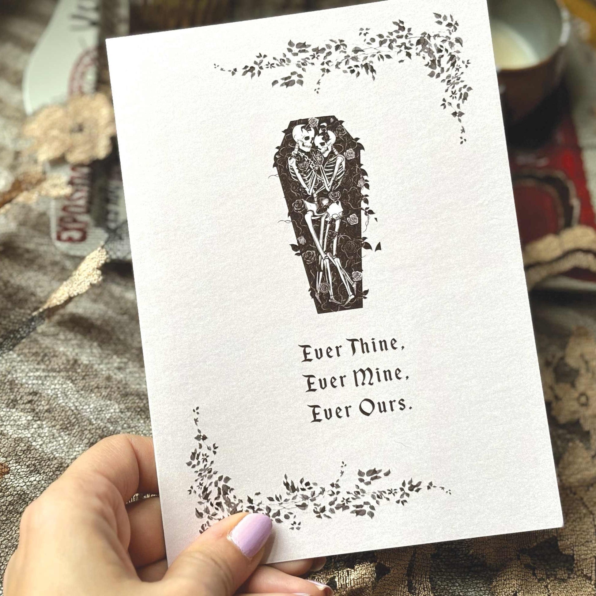 A greeting card featuring a black ink illustration of a skeleton in a coffin, surrounded by floral vines and the text 'Ever Thine. Ever Mine. Ever Ours.