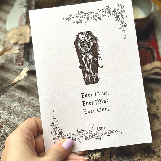 A greeting card featuring a black ink illustration of a skeleton in a coffin, surrounded by floral vines and the text 'Ever Thine. Ever Mine. Ever Ours.