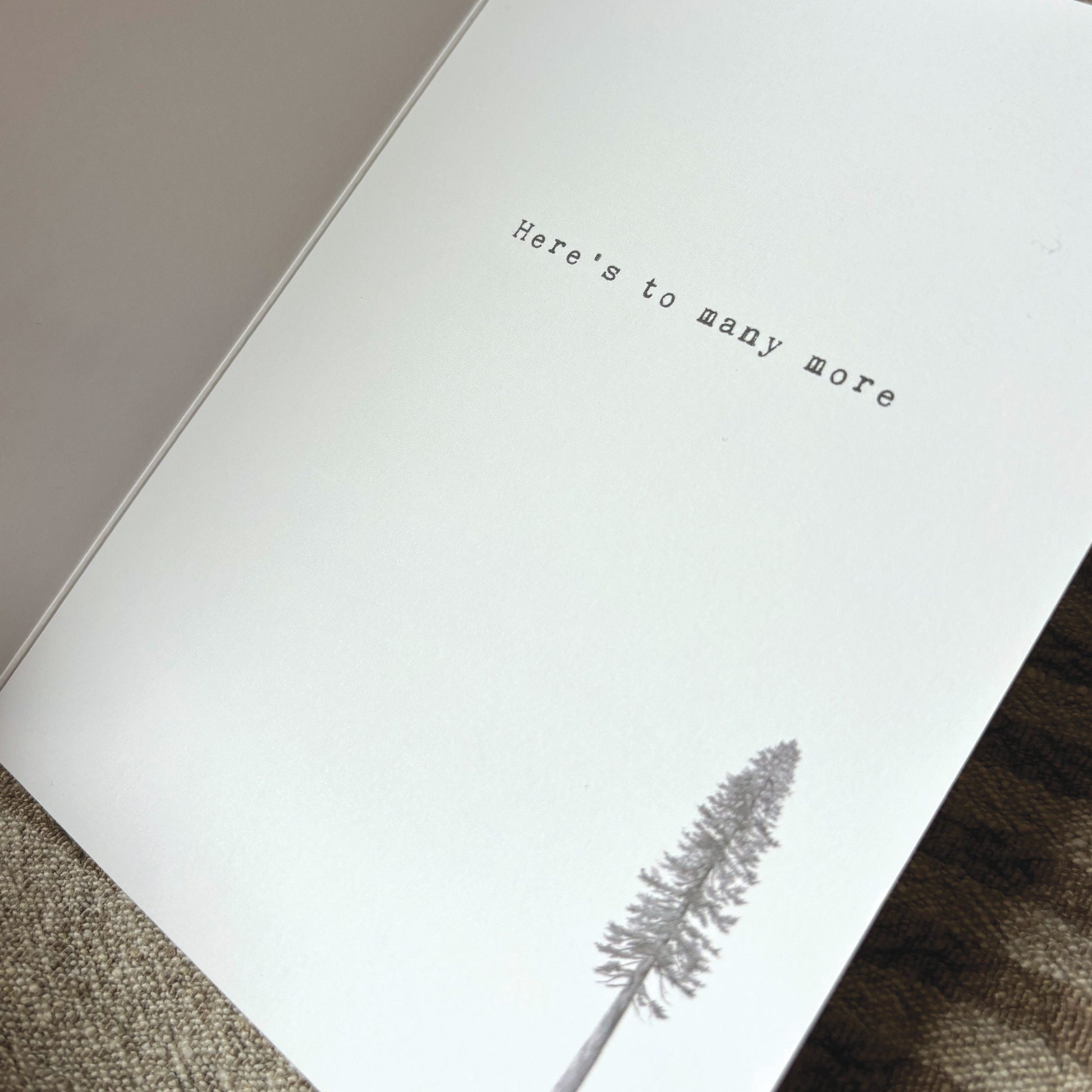 Inside of greeting card which says "here's to many more" with an illustration of a pine tree