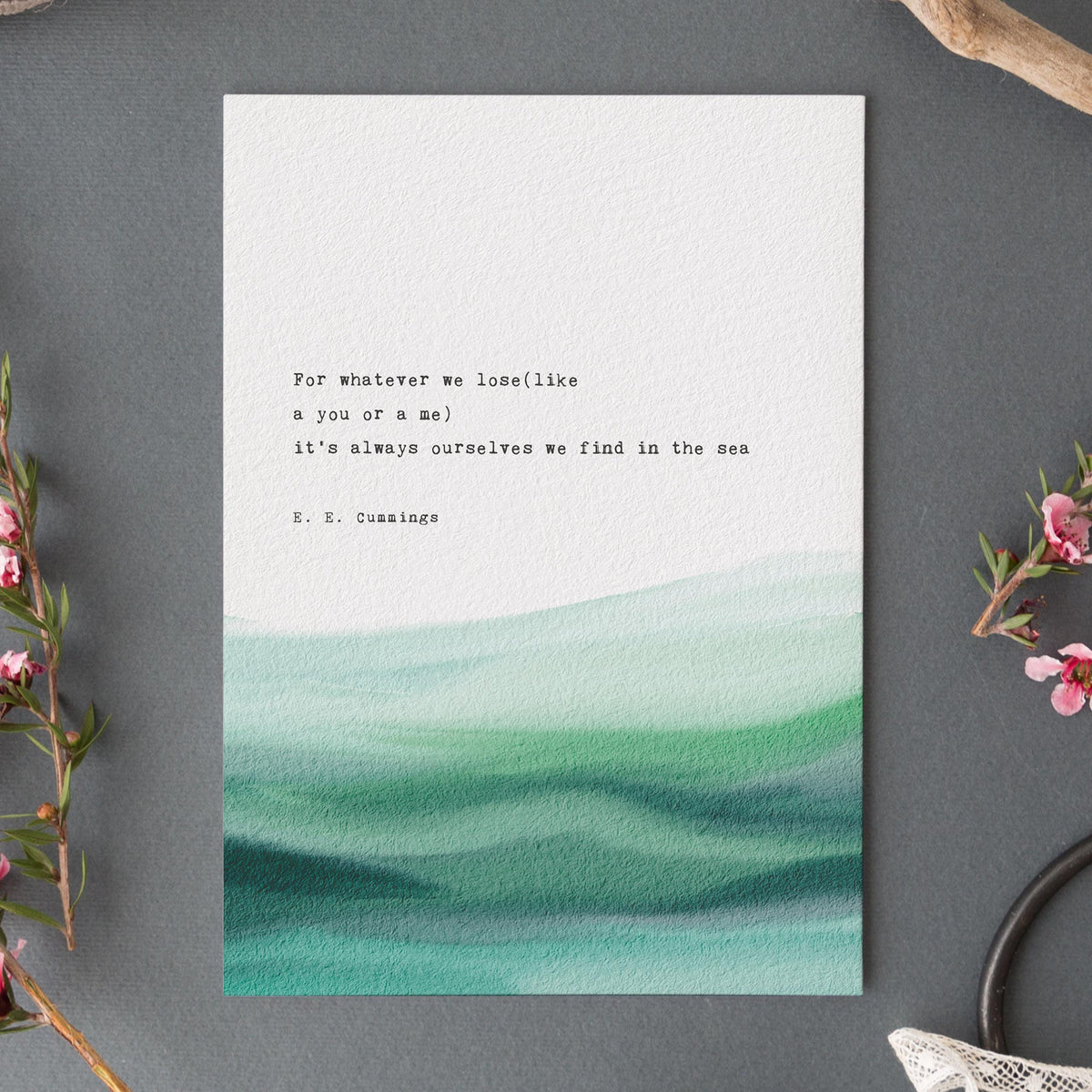 An art print featuring a quote by e.e. cummings on a soft watercolor background which says "for whatever we lose(like a you or a me) it's always ourselves we find in the sea"