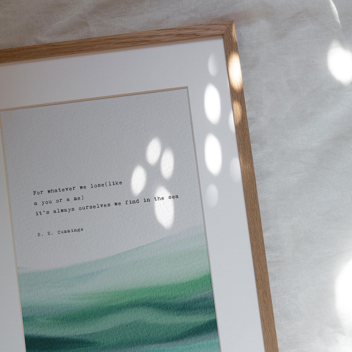 An art print featuring a quote by e.e. cummings on a soft watercolor background which says "for whatever we lose(like a you or a me) it's always ourselves we find in the sea"