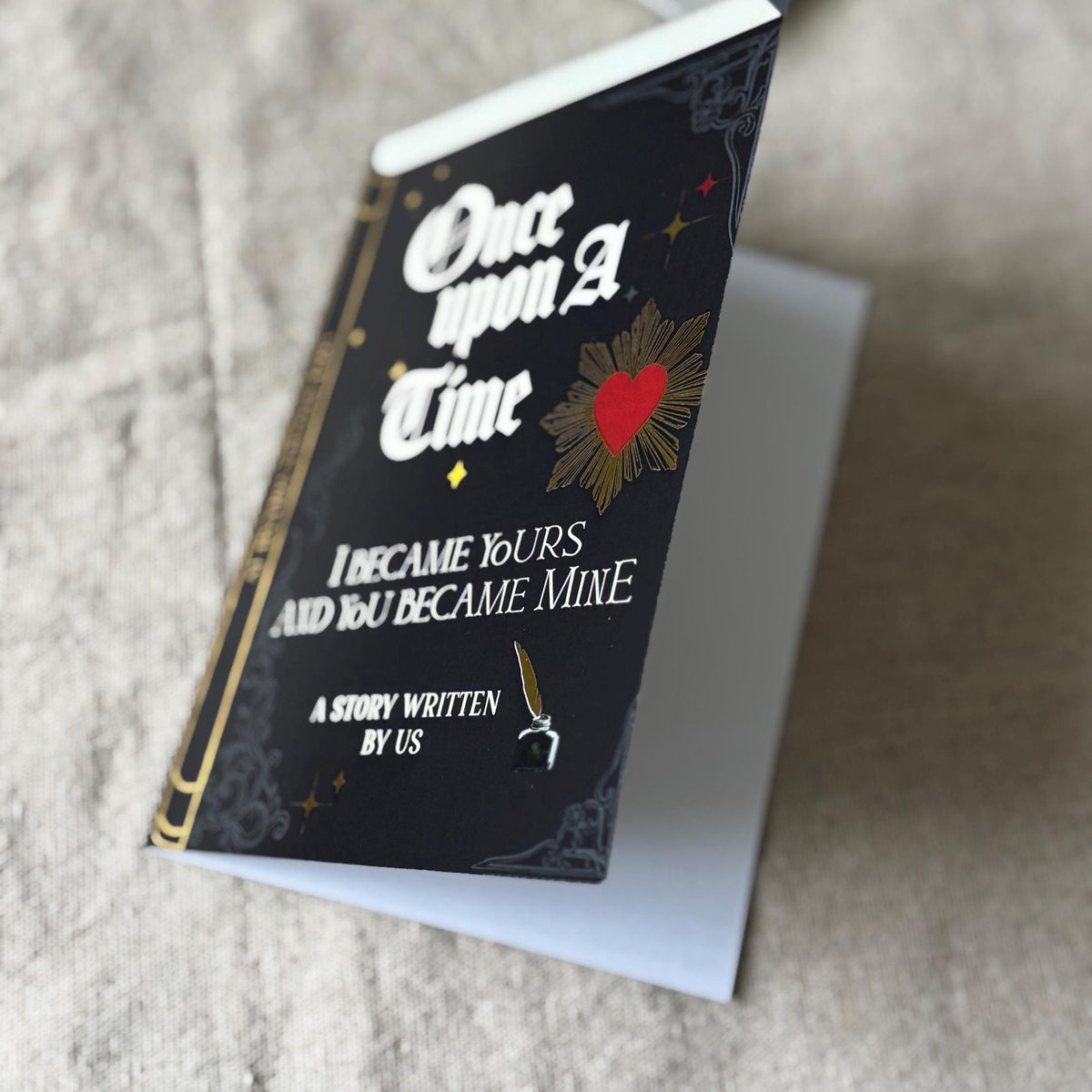 A greeting card that looks like a black hardcover book titled 'Once upon a Time I Became Yours and You Became Mine' with gothic white lettering and gold decorative details.