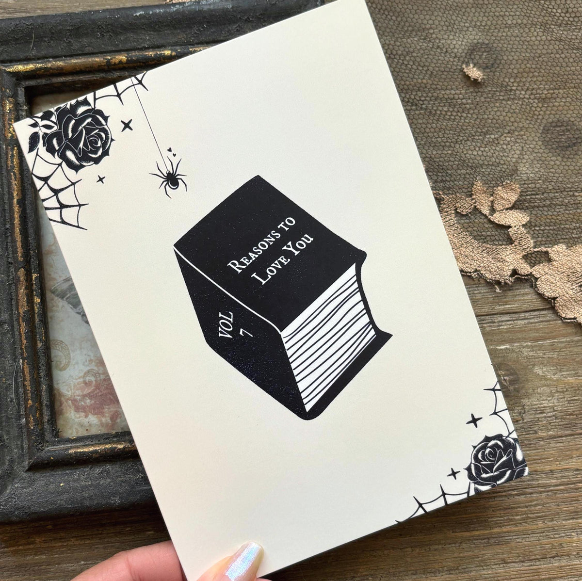 A white greeting card featuring a black illustration of a book titled 'Reasons to Love You' with spiderwebs, roses, and stars