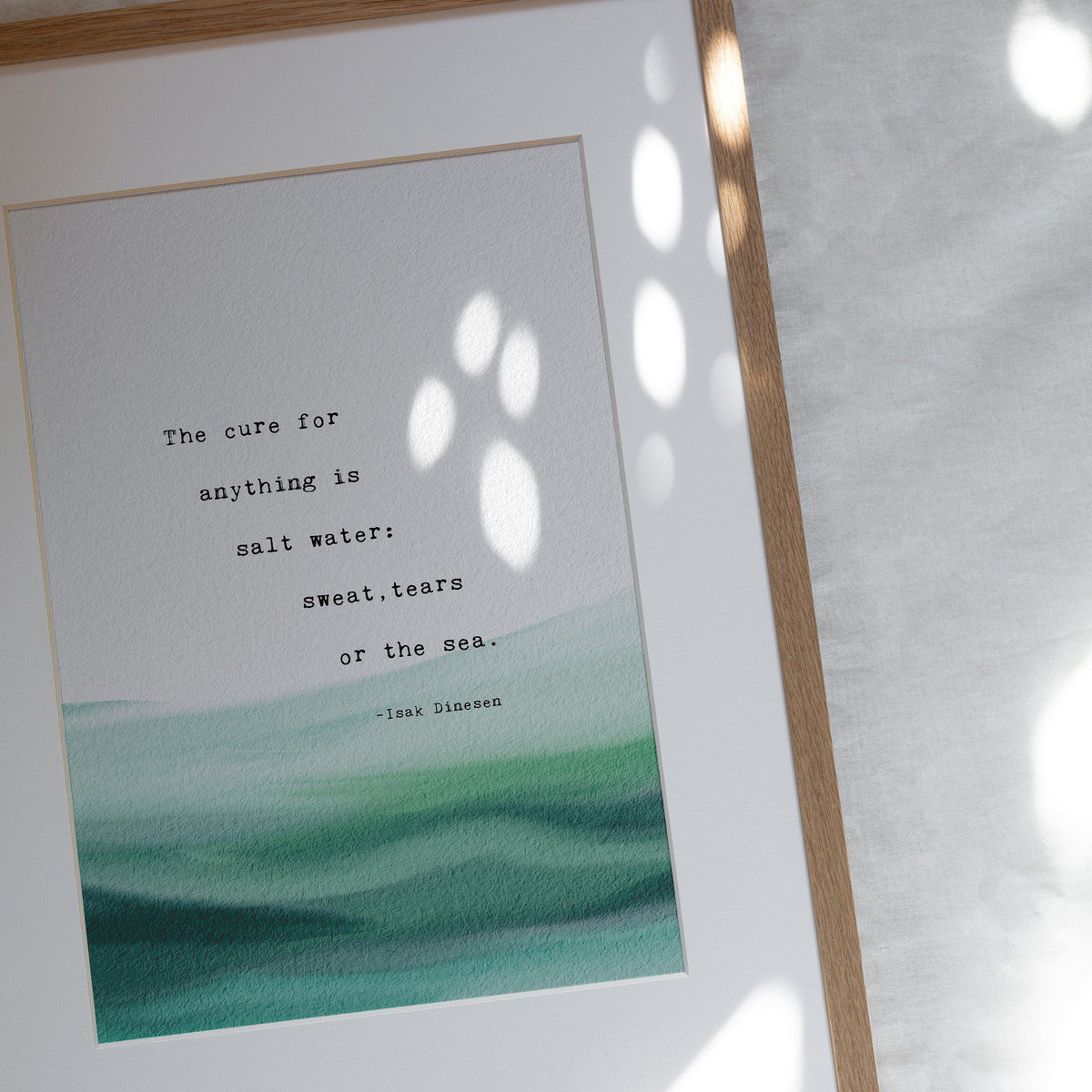 A typographic art print featuring a quote by Isaac Dinesen over a watercolor ocean wave design in shades of teal and green on a white background. "The cure for anything is salt water: sweat, tears or the sea"