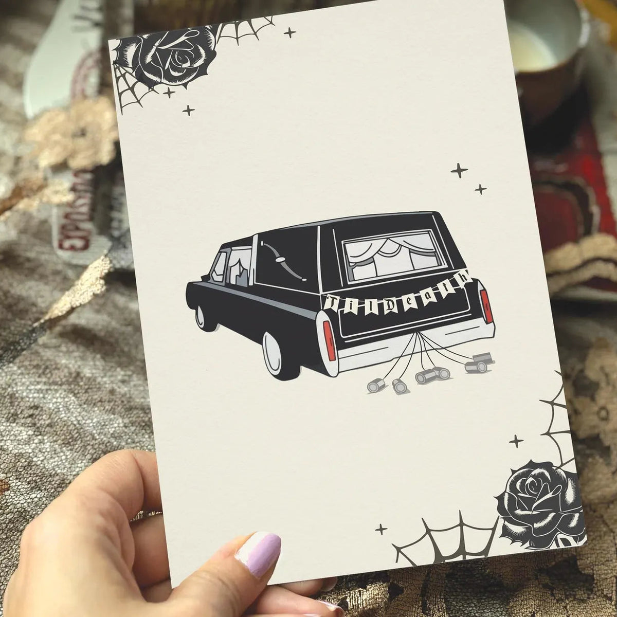 A cream-colored greeting card featuring a black-and-white illustration of a hearse with 'TIl Death' on the back, decorated with spiderwebs and roses. 