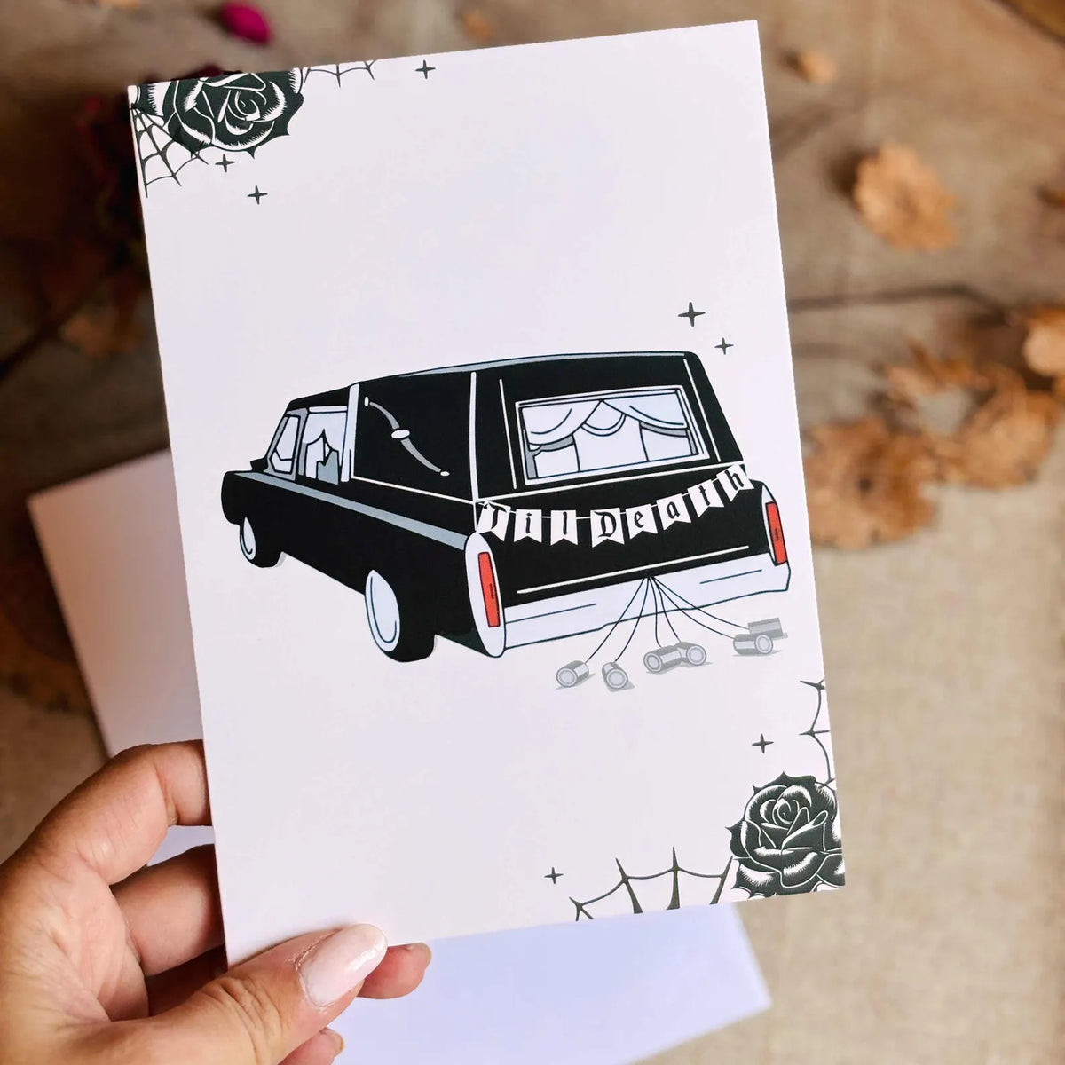 A cream-colored greeting card featuring a black-and-white illustration of a hearse with 'TIl Death' on the back, decorated with spiderwebs and roses. 