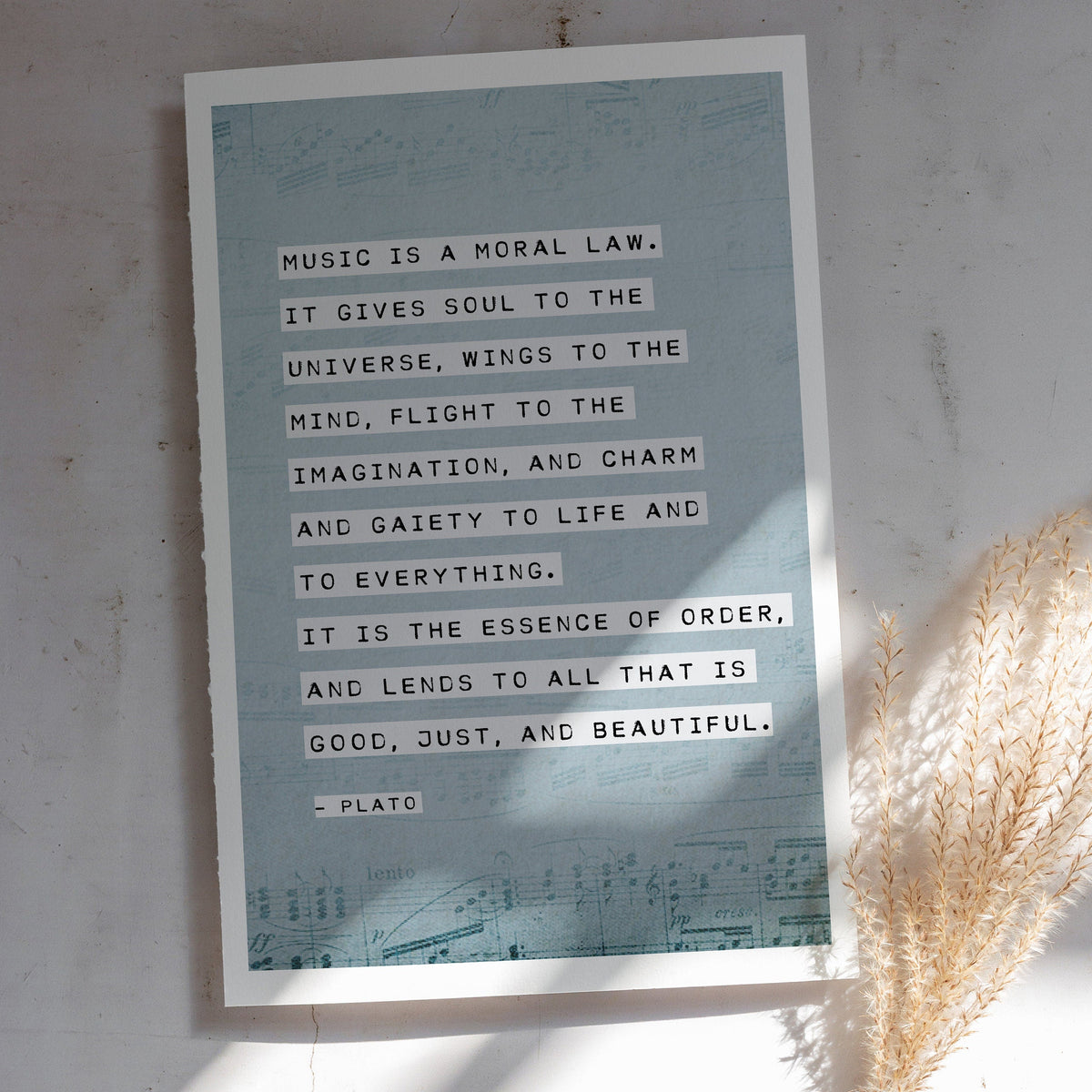 An art print of sheet music on a pale blue background with the quote by Plato "Music is a moral law, it gives soul to the universe, wings to the mind..."